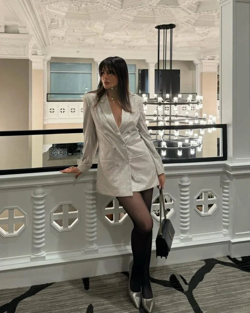 Woman in silver blazer dress with metallic heels leaning on railing indoors, New Year’s Eve outfit.
