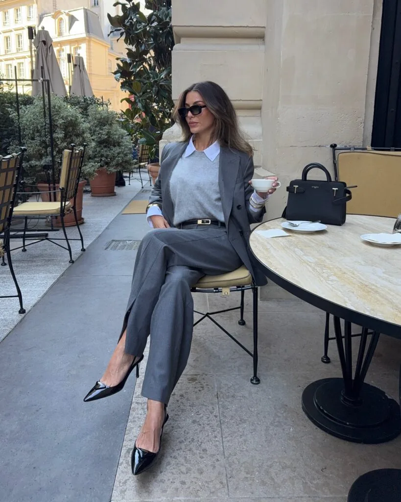 Grey suit with light grey sweater and white collar, styled with black heels at a café table.
