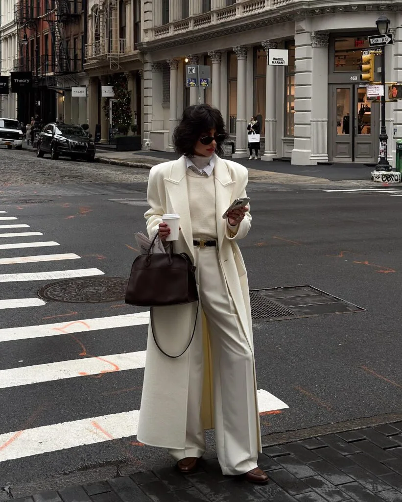 White long coat with cream sweater and wide-leg trousers, holding coffee and a brown bag on a crosswalk.

