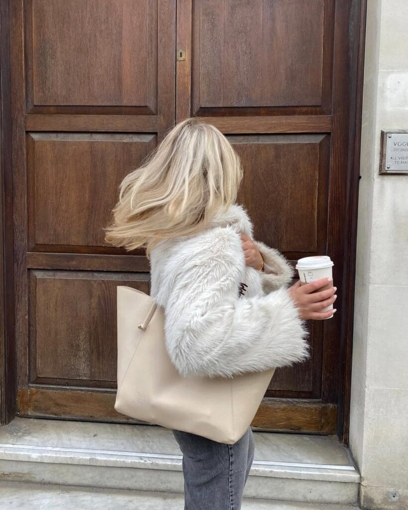 Blonde woman in white fuzzy jacket, Cuddly Teddy Layers trend, carrying a cream tote and coffee cup