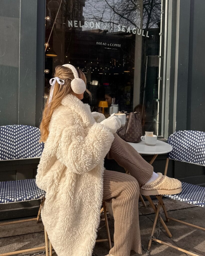 Seated woman in ivory teddy coat and earmuffs, Cuddly Teddy Layers trend, outdoor café winter scene.