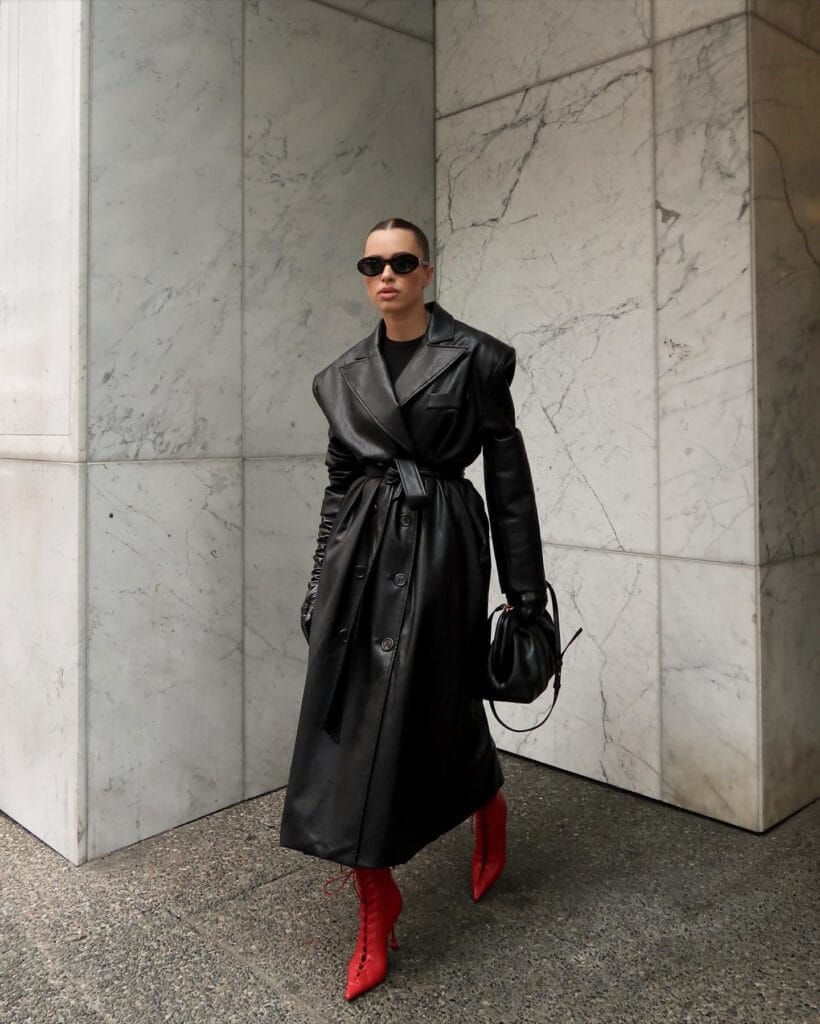 Woman in long black leather trench coat and red boots, Pop-of-Red trend, marble wall backdrop.