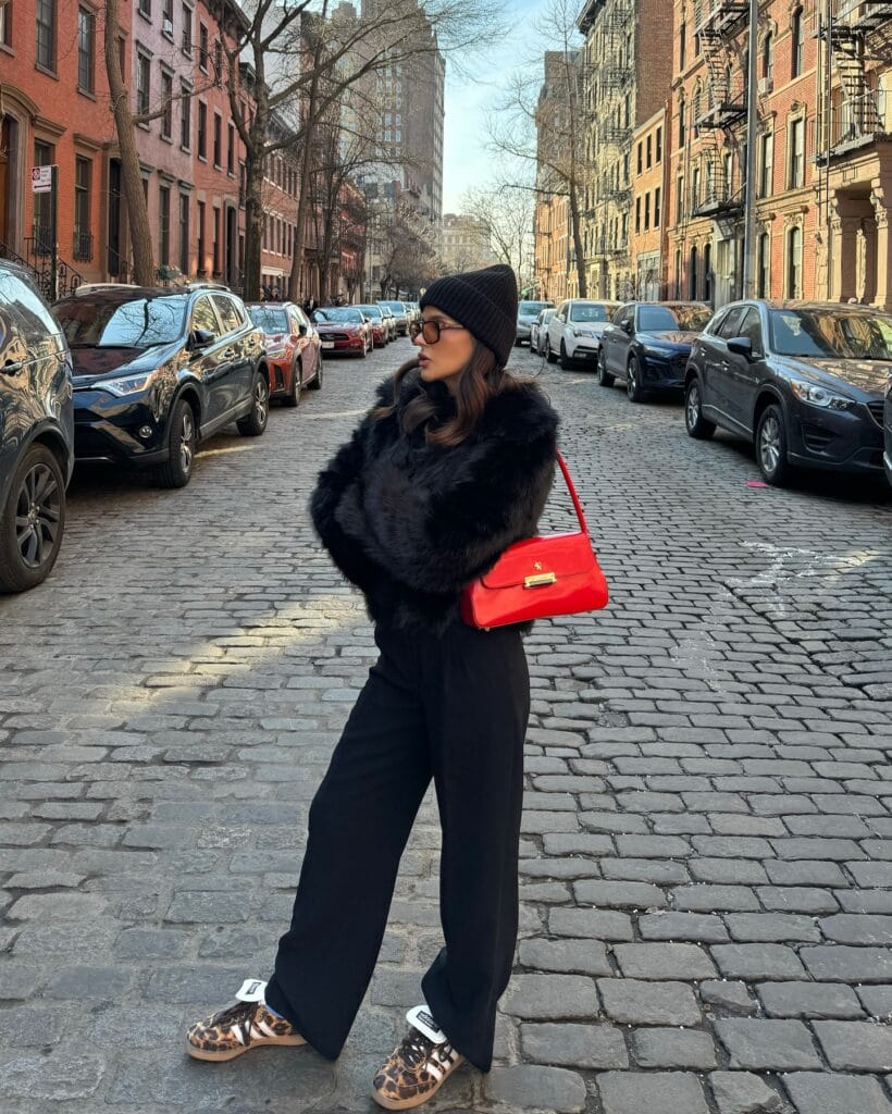 Black faux fur jacket with red shoulder bag, Pop-of-Red trend, city street style with sneakers and beanie.