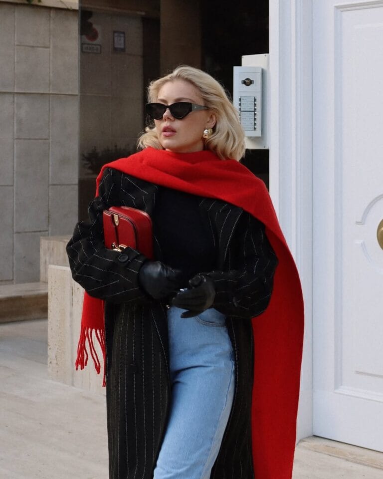 Blonde woman in black coat and red scarf, Pop-of-Red trend, holding matching red bag and wearing sunglasses.