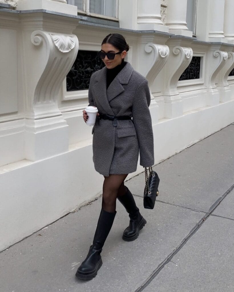 Woman in a gray belted coat with tall black boots, Belted & Cinched trend, city sidewalk style.