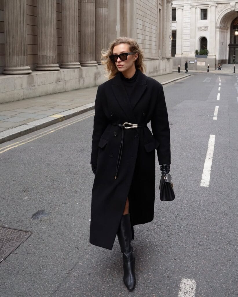 Long black coat with gold sculptural belt, Belted & Cinched trend, city street, tall black boots and sunglasses.