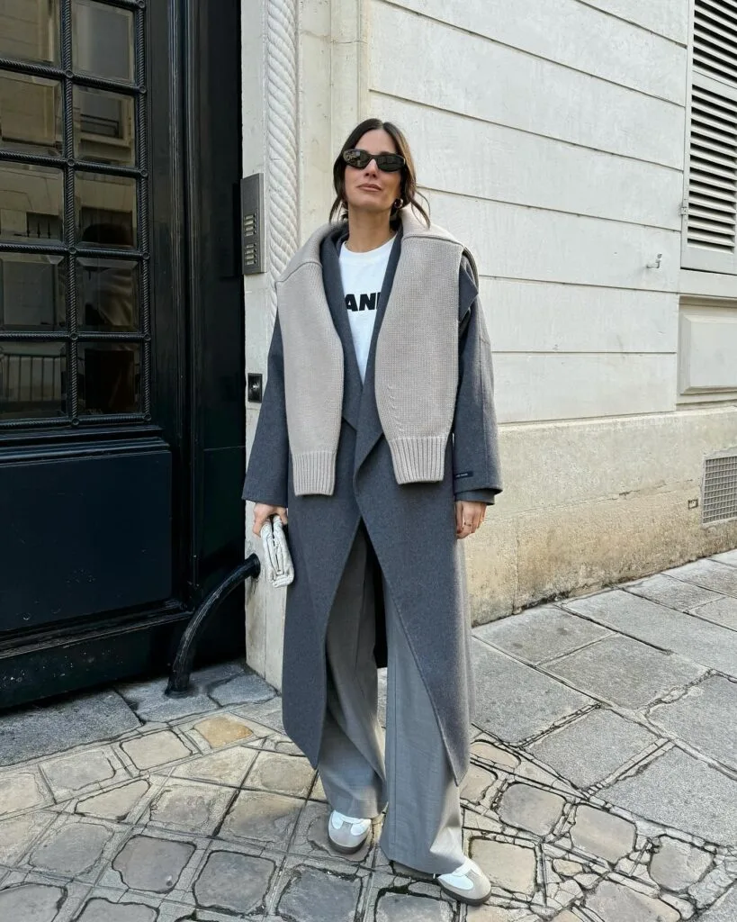 Grey long coat and trousers with sweater draped over shoulders and white tee—minimal casual winter outfits.
