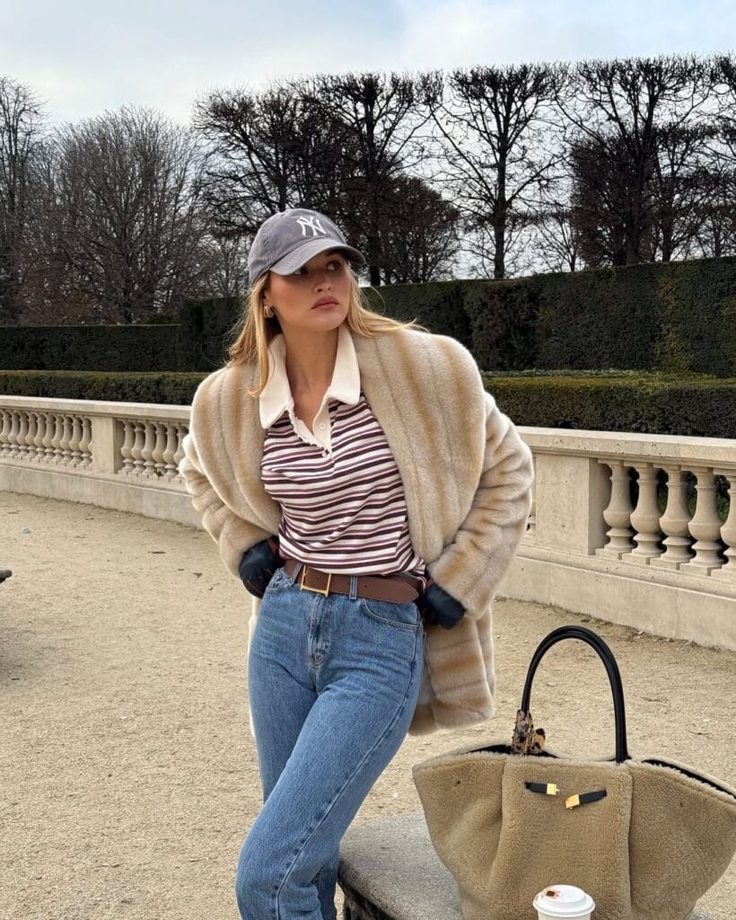 winter paris outfit with faux fur jacket, striped polo, blue jeans, and baseball cap

