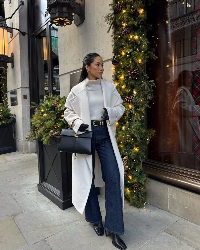 woman in ivory coat, white turtleneck, dark jeans, black gloves, standing by Christmas decor