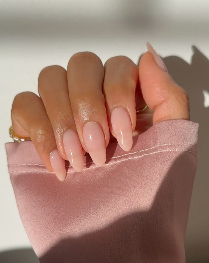 sheer pink almond nails with glossy finish, gold rings, pink fabric background