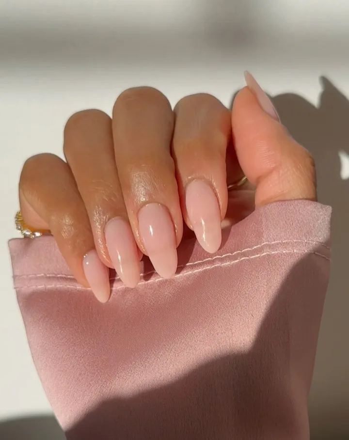 sheer pink almond nails with glossy finish, gold rings, pink fabric background