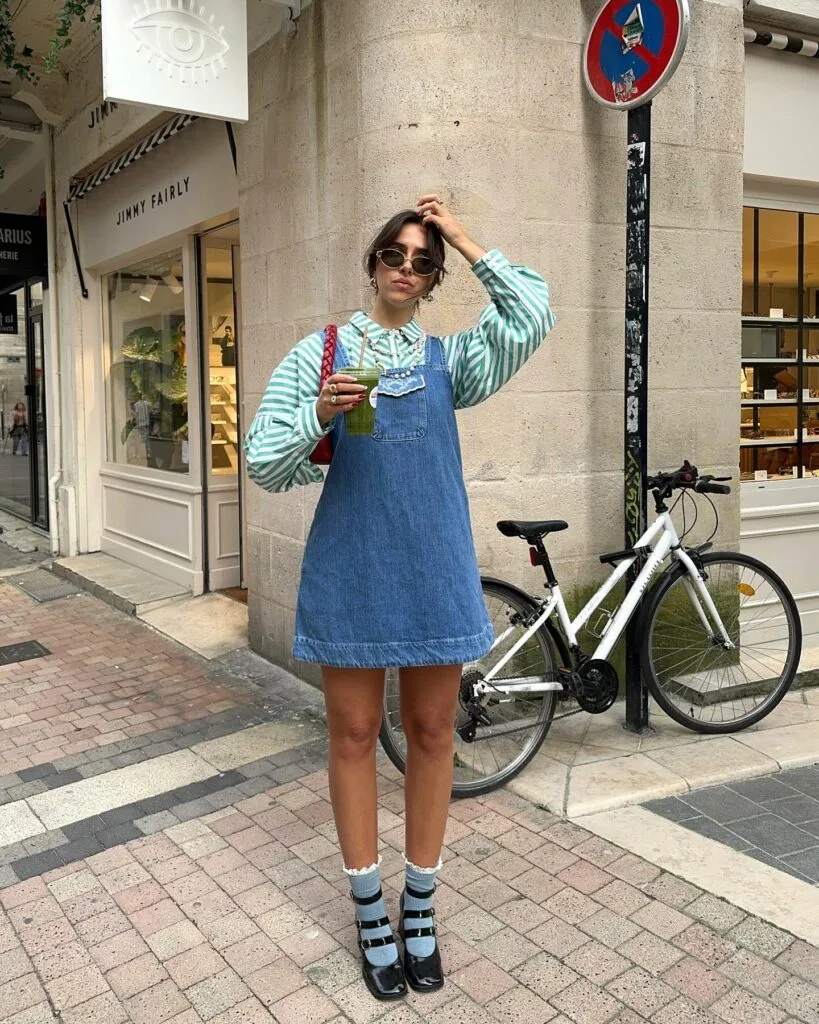 pring fashion trends 2026, denim pinafore dress over green striped button-down with Mary Jane shoes
