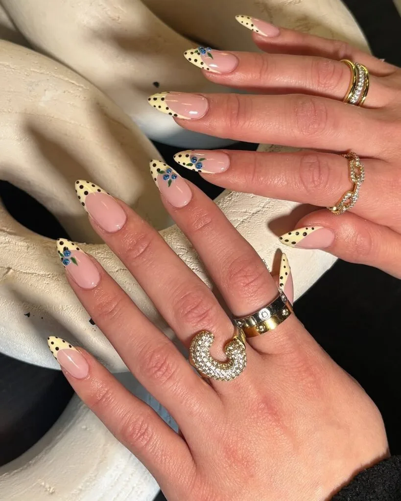 may nails, almond nude nails with butter yellow polka dot French tips and hand-painted blue floral accents
