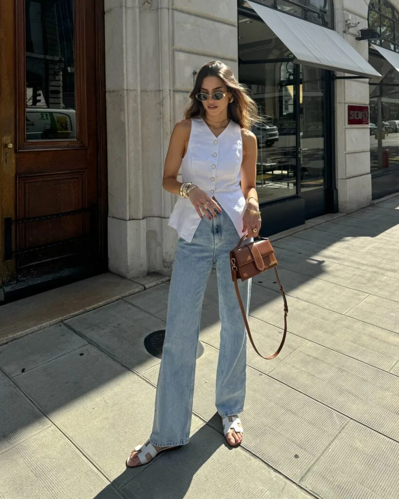 june outfit with white sleeveless button-front top, light wash wide-leg jeans, white flat sandals, and cognac bag
