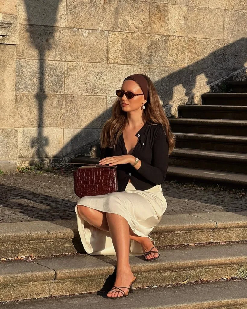 summer outfit with black button-front cardigan, cream midi skirt, strappy heeled sandals, and burgundy croc clutch
