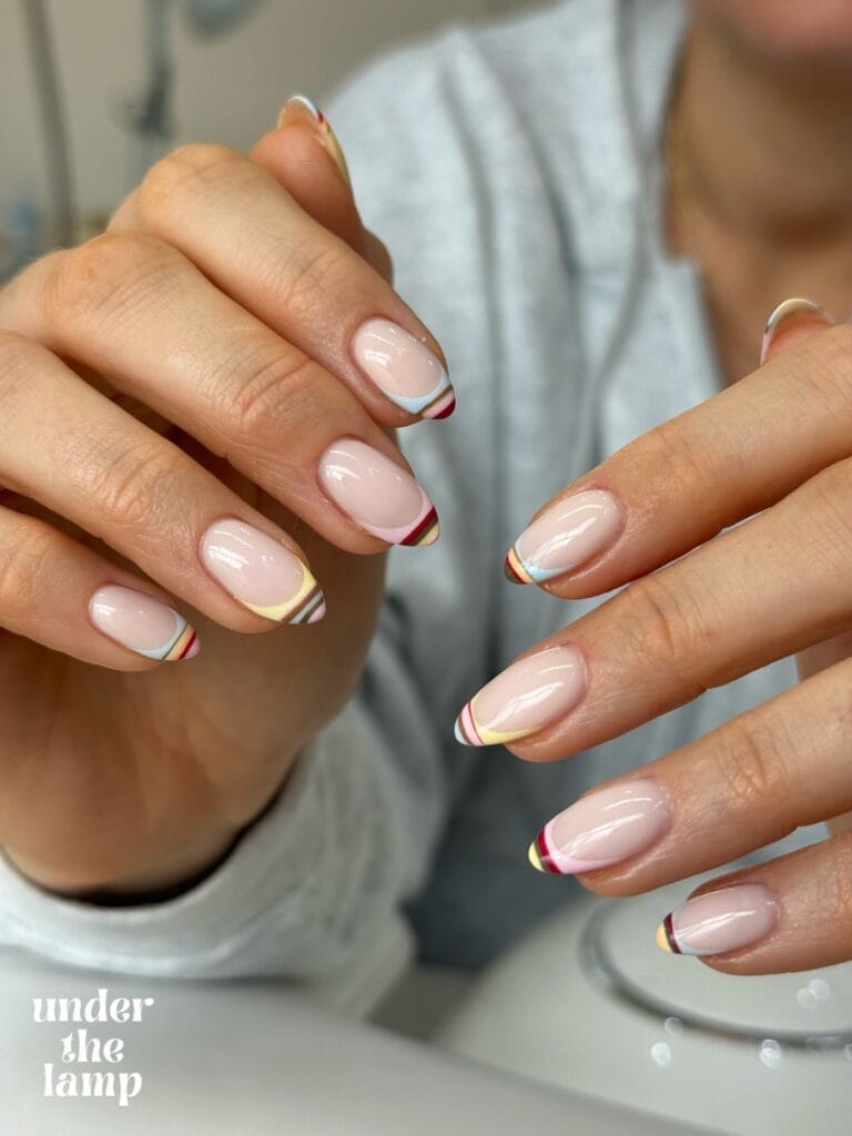 June nail ideas, sheer nude nails with thin rainbow stripe French tips in red yellow blue and white, text "under the lamp"
