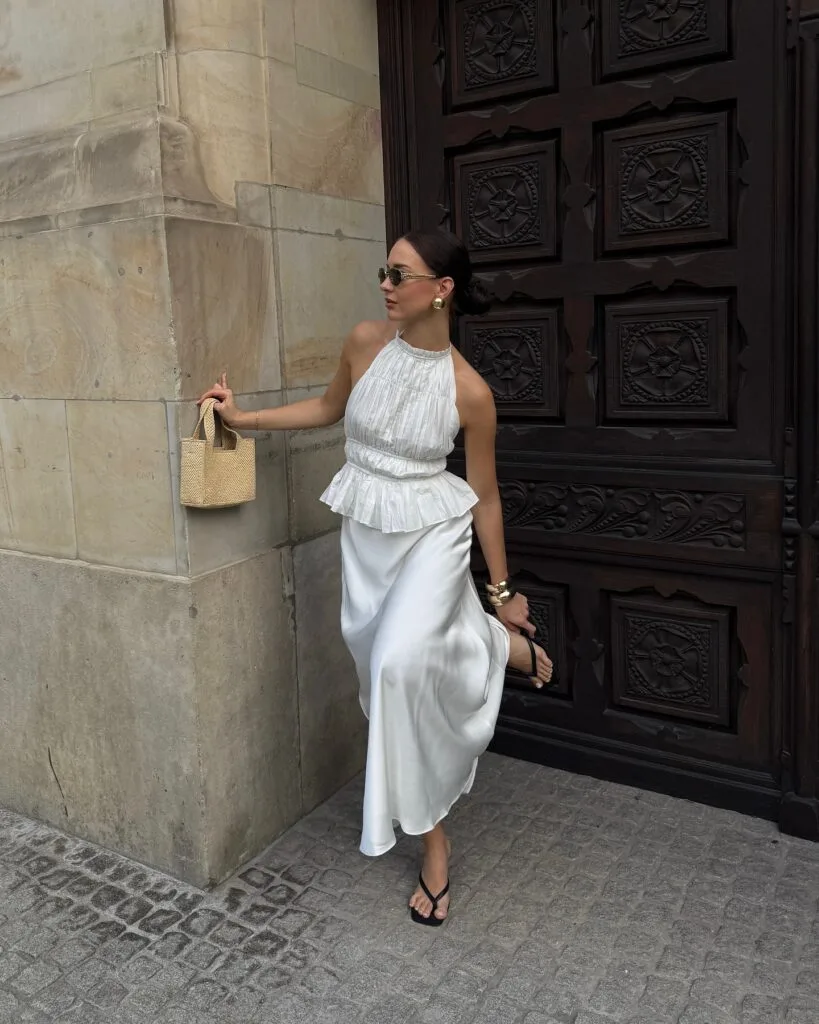 summer outfit with white pleated halter top, white satin midi skirt, black flip-flop sandals, and woven bag

