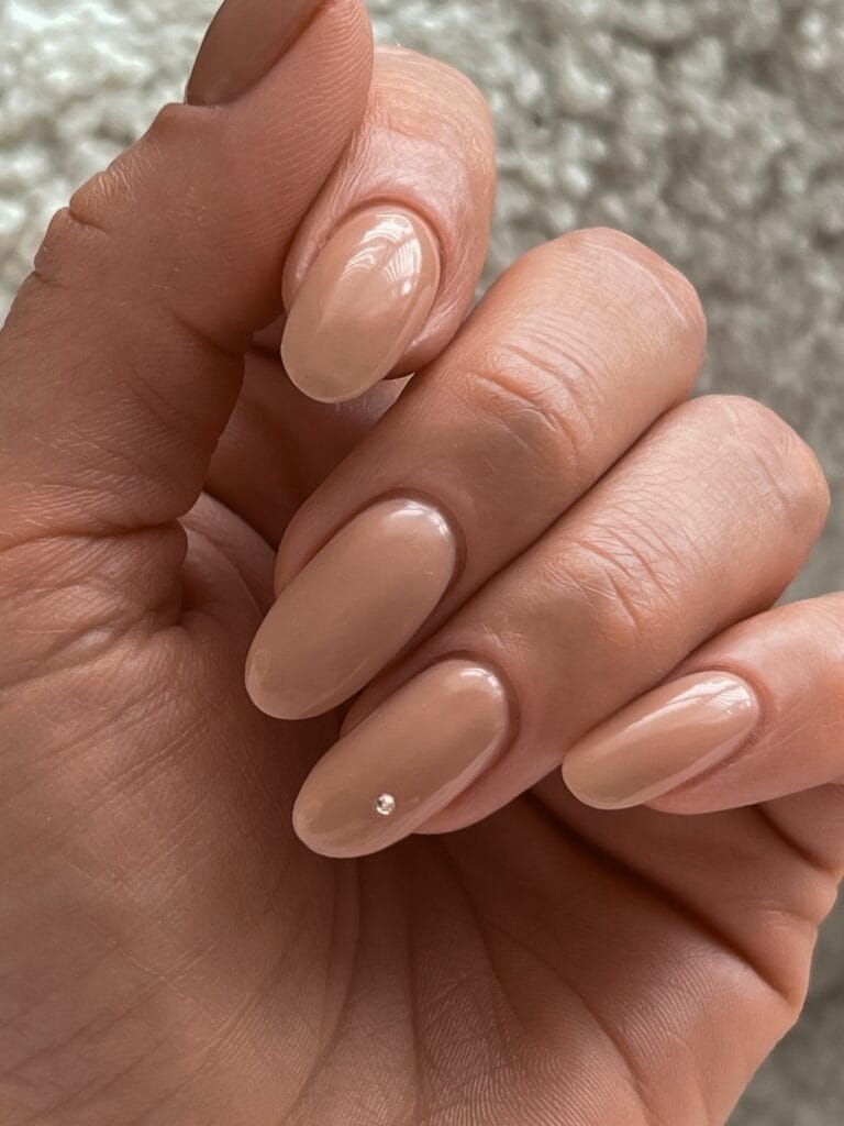Glossy beige nude nails with an almond shape and one tiny crystal accent.
