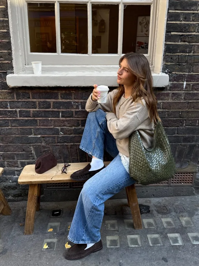 Fall fashion outfit with beige knit sweater, lace-trim top, wide-leg jeans, loafers, and green woven bag.
