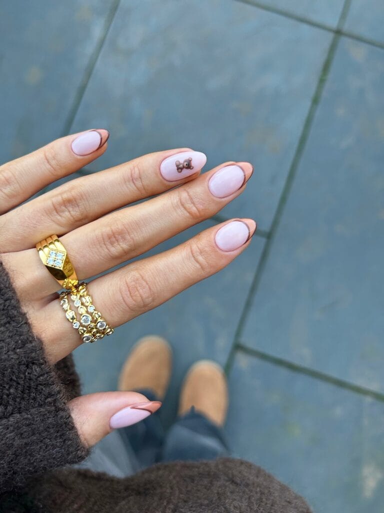 Milky autumn nails with thin mocha curves, warm brown corner details, and one tiny teddy bear accent.