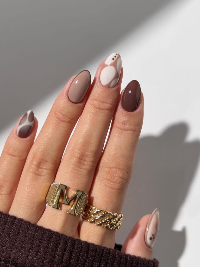 Neutral fall nails in cream, taupe, and chocolate with abstract curved designs and tiny brown dots.