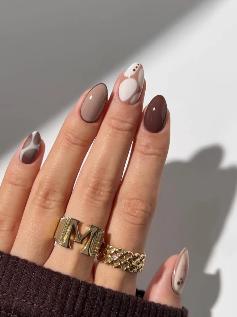 Neutral fall nails in cream, taupe, and chocolate with abstract curved designs and tiny brown dots.