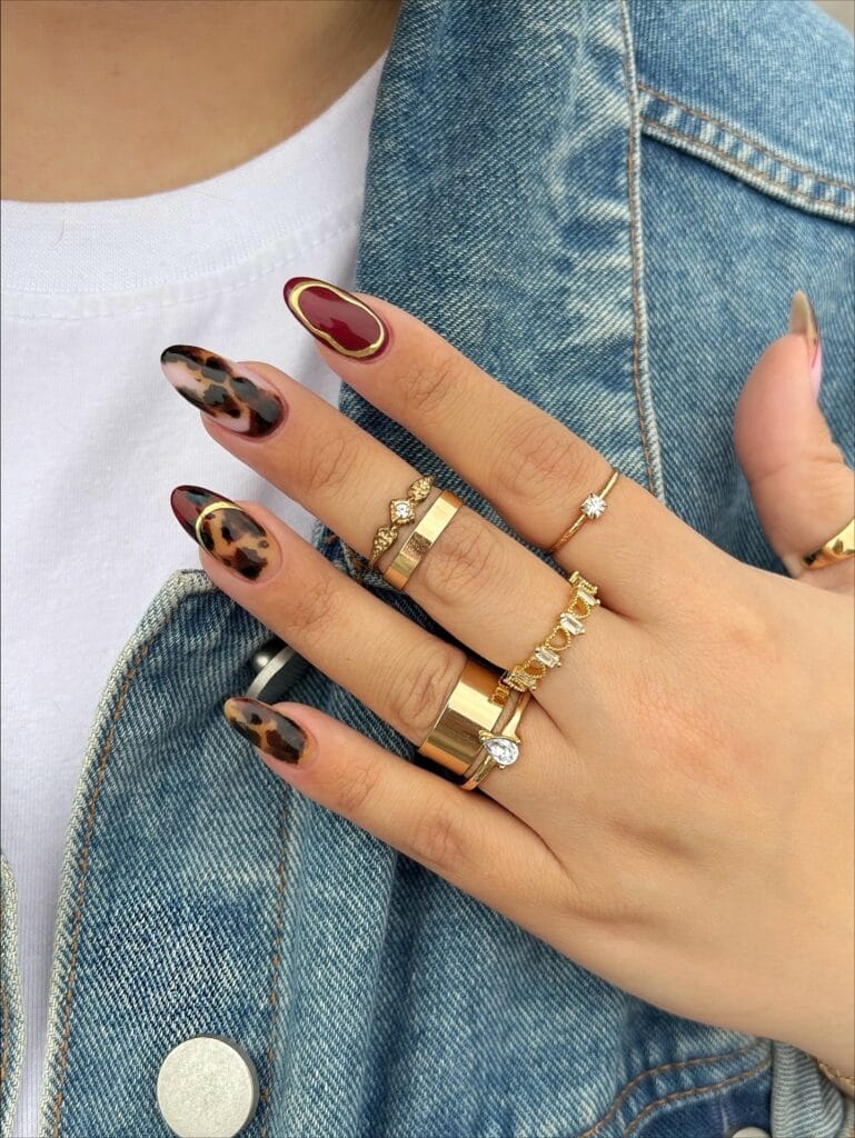 Almond tortoiseshell nails with burgundy accents and thin metallic gold outlines.
