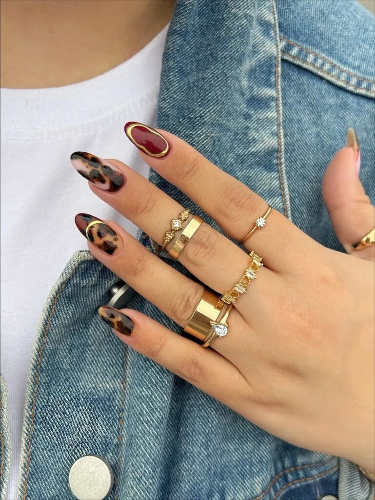 Almond tortoiseshell nails with burgundy accents and thin metallic gold outlines.
