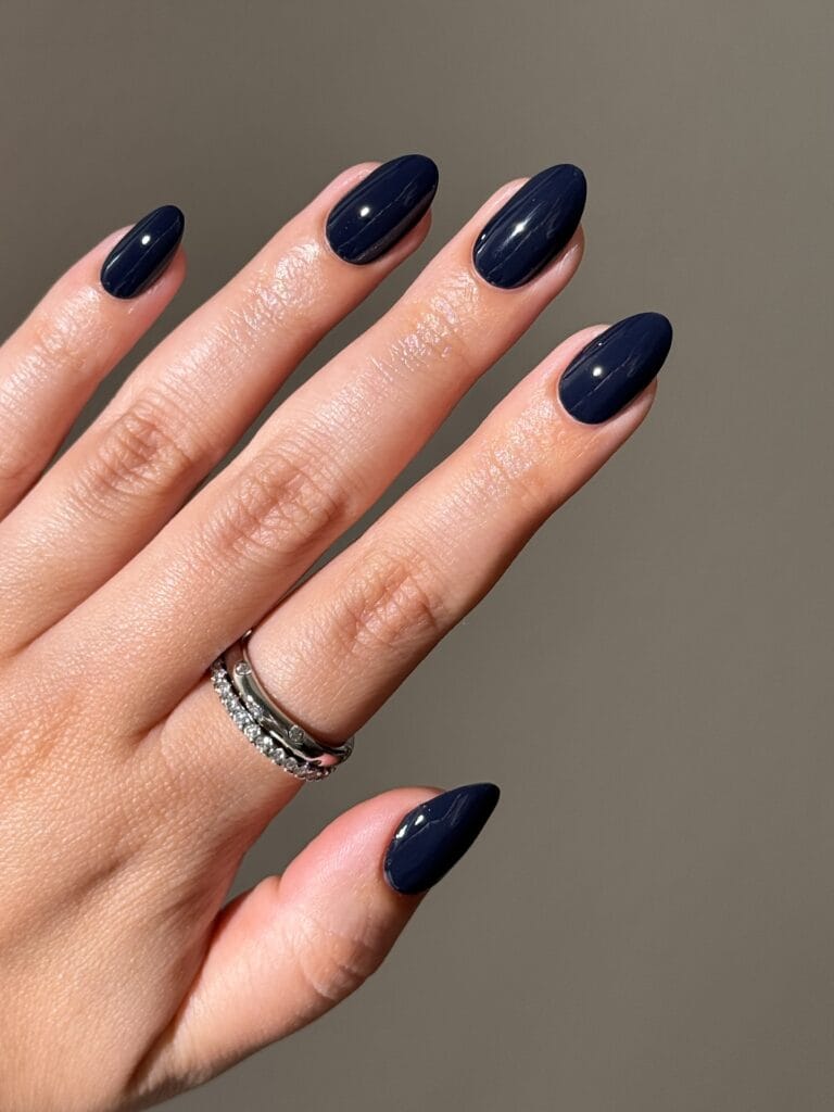 Glossy midnight blue almond nails in sunlight with silver rings, a fall nails look.