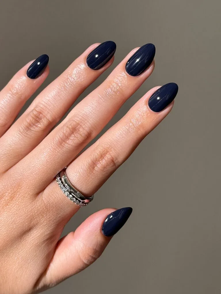 Glossy midnight blue almond nails in sunlight with silver rings, a fall nails look.