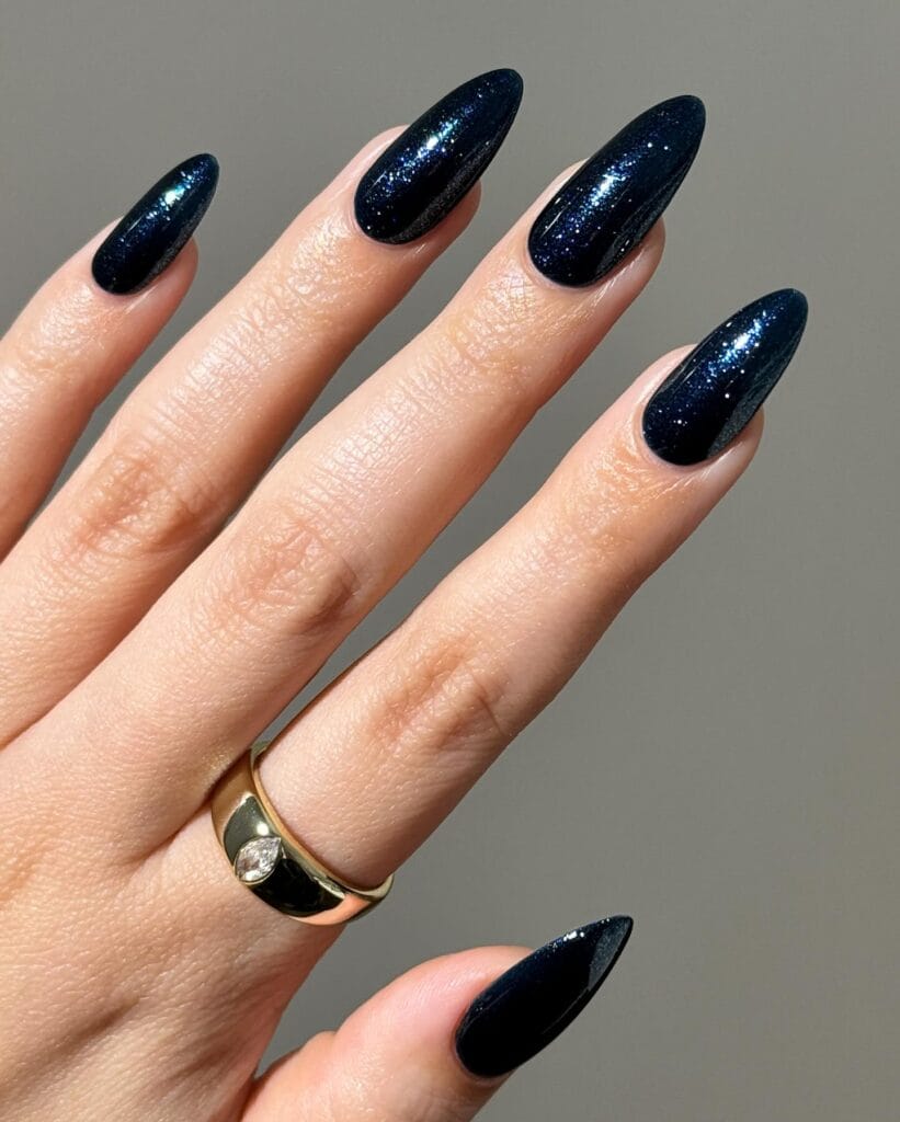 Shimmery midnight blue almond nails with fine sparkle and a gold ring, an autumn nails idea.
