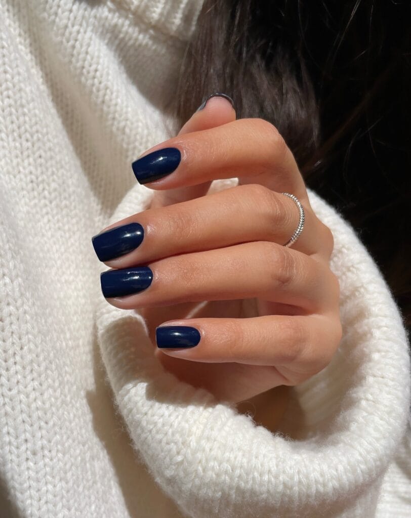 Short square midnight blue nails with a glossy finish against a white knit sweater, fall nail colors.