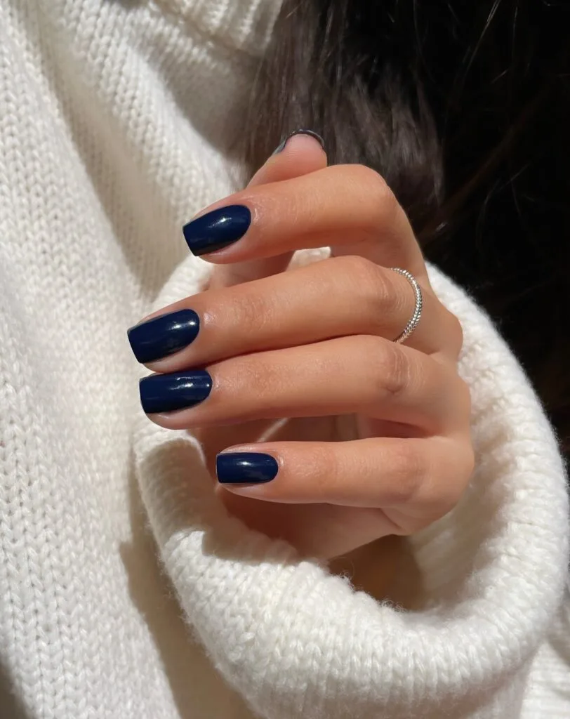Short square midnight blue nails with a glossy finish against a white knit sweater, fall nail colors.