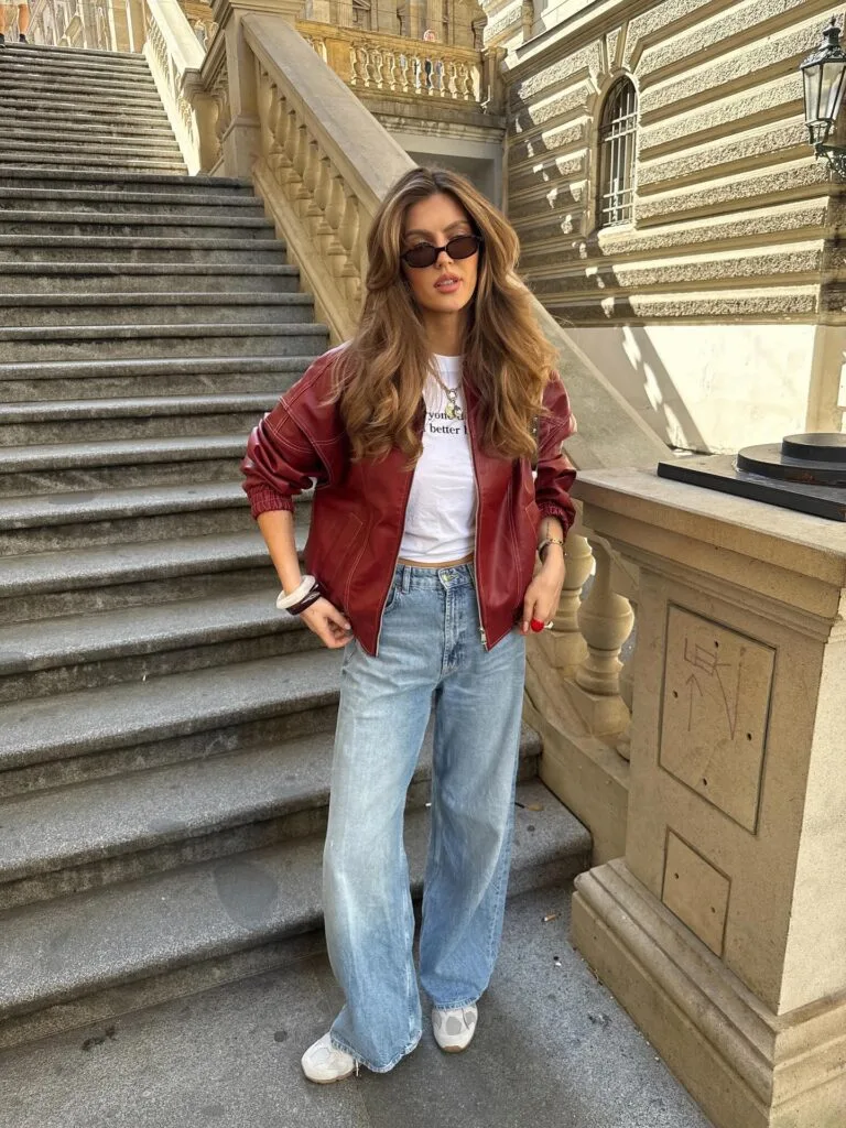 Fall outfit with burgundy leather bomber jacket, white tee, wide-leg jeans, and sneakers.
