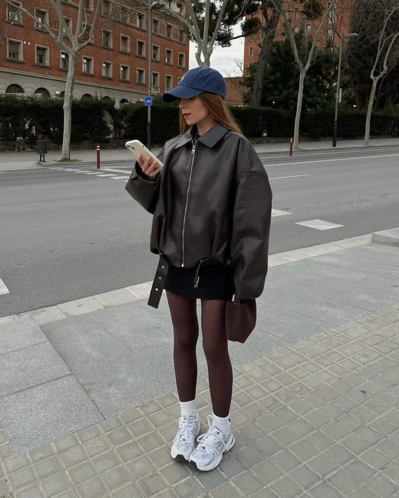 Woman in a brown leather jacket, mini skirt, burgundy tights, and sneakers in an autumn outfit.