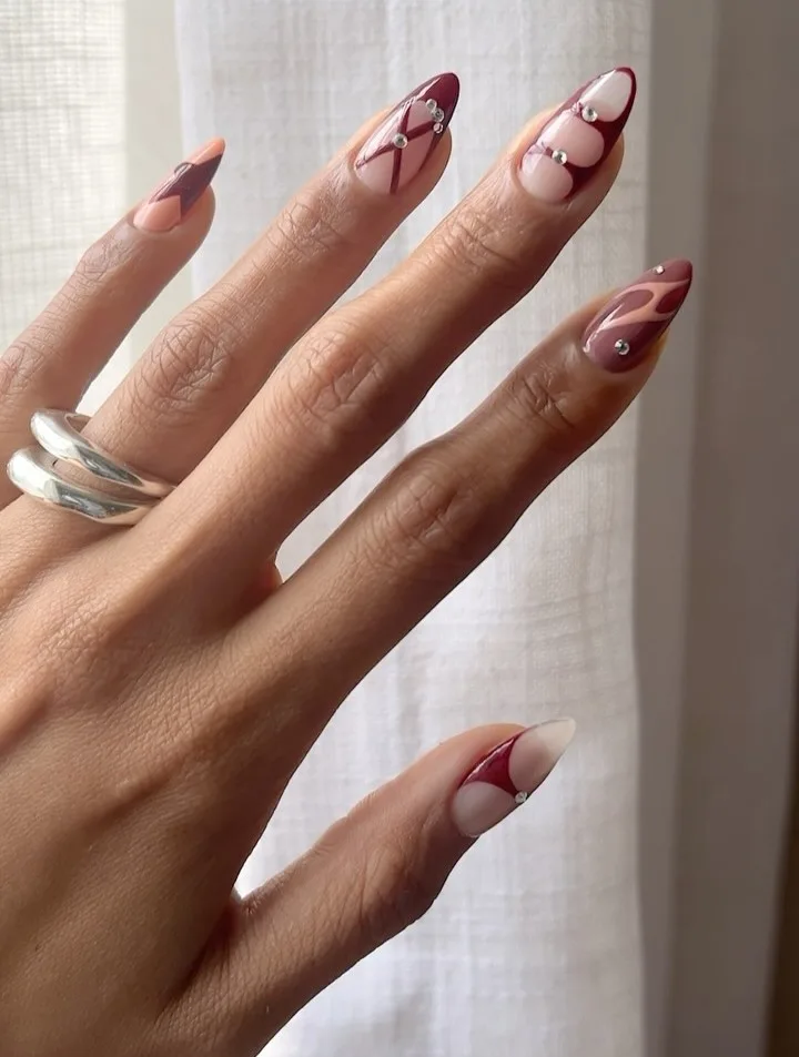 Almond early fall nails with burgundy graphic swirls, negative space, and silver gem accents.