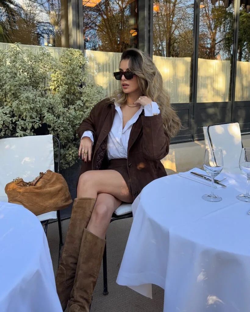 Brown suede jacket, knee-high suede boots, and tan bag in a fall 2026 outfit at an outdoor café.