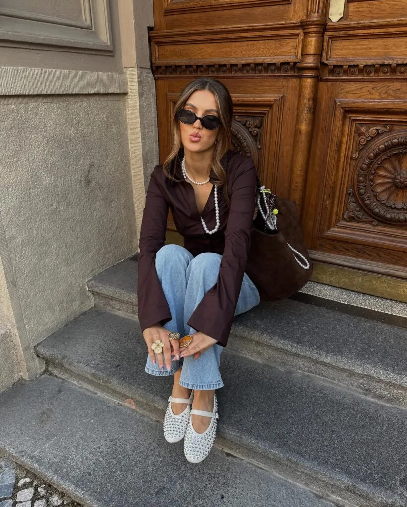 Autumn outfit with brown sheer blouse, light blue jeans, embellished Mary Jane flats, and brown bag.