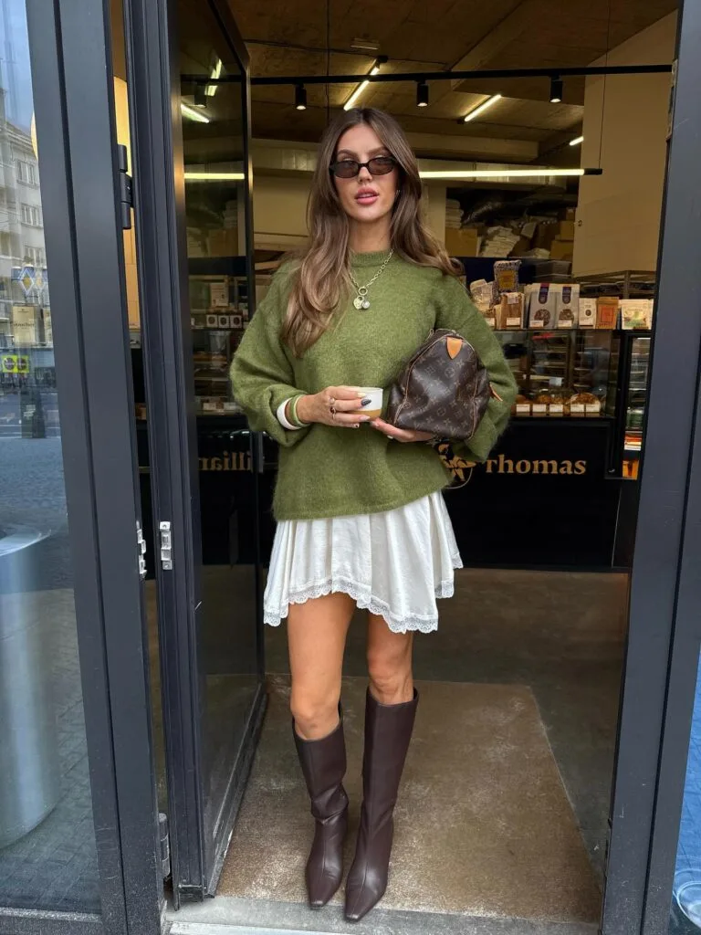 Early fall outfit with green sweater, white mini dress, and brown knee-high boots.