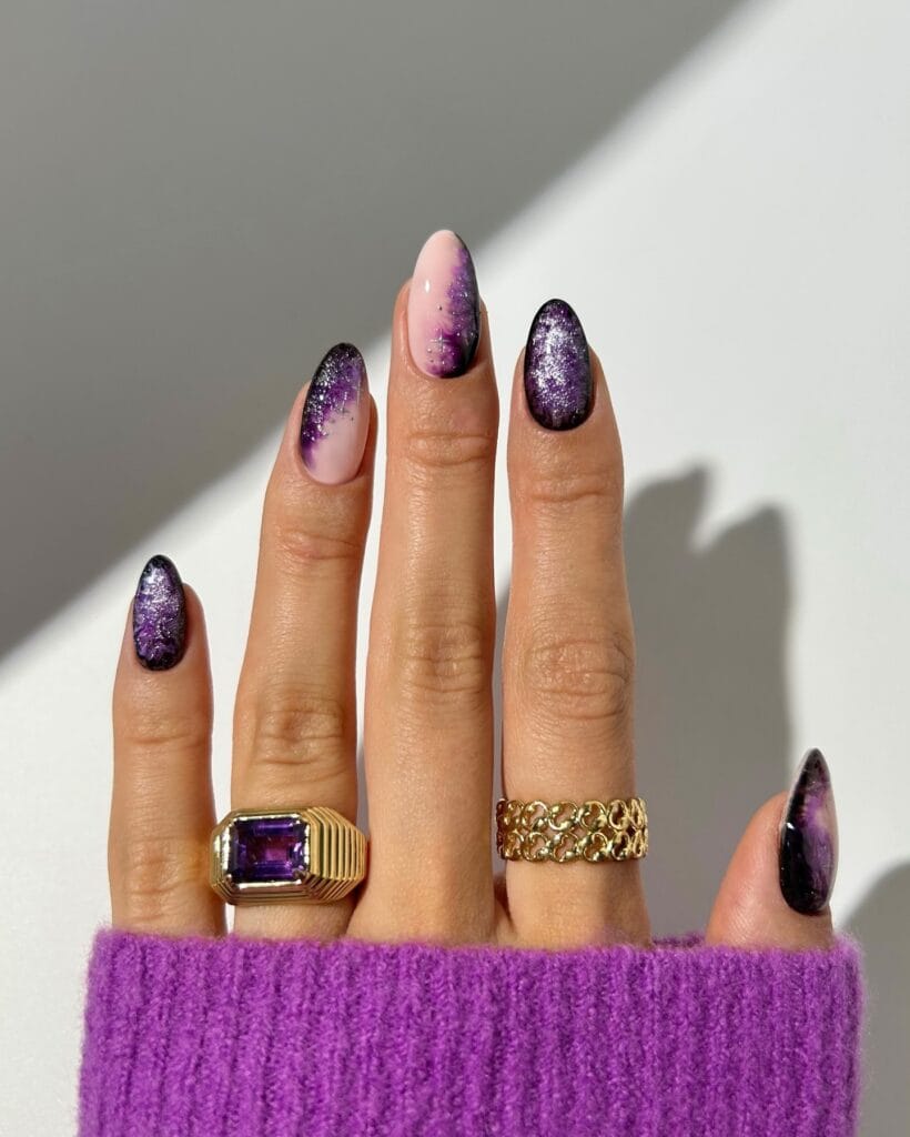 Purple and black witchy nails with smoky swirls, silver flecks, and sheer pink accents on almond shapes.