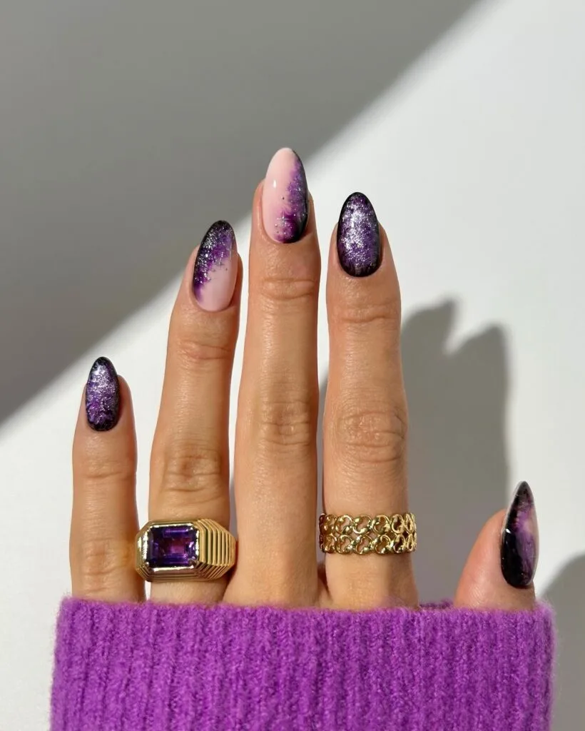 Purple and black witchy nails with smoky swirls, silver flecks, and sheer pink accents on almond shapes.