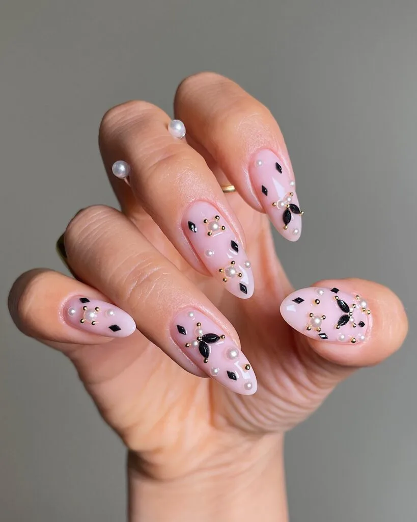 Pale pink witchy nails with black diamond-shaped gems, white pearls, and tiny gold studs on long almond shapes.