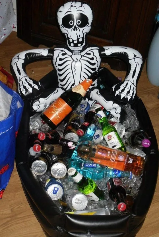 Halloween drink tub with a skeleton reclining in ice among assorted bottles and cans
