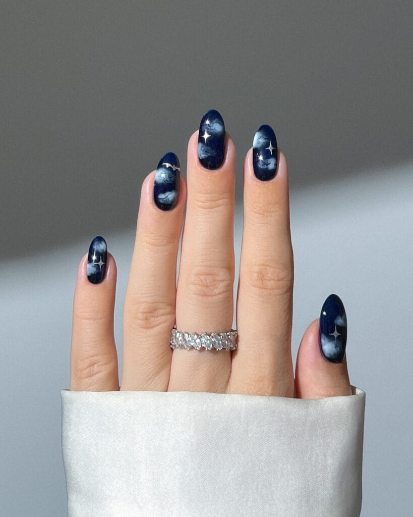 Navy blue witchy nails with wispy pale clouds, silver stars, and a tiny bat on oval shapes.