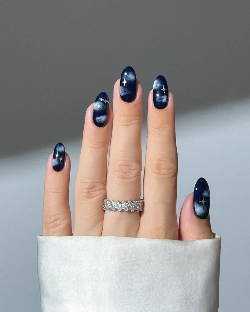 Navy blue witchy nails with wispy pale clouds, silver stars, and a tiny bat on oval shapes.
