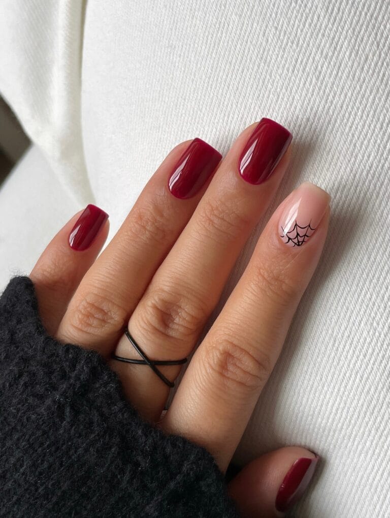 short Halloween nails with glossy berry red polish and one nude accent nail with black spiderweb art