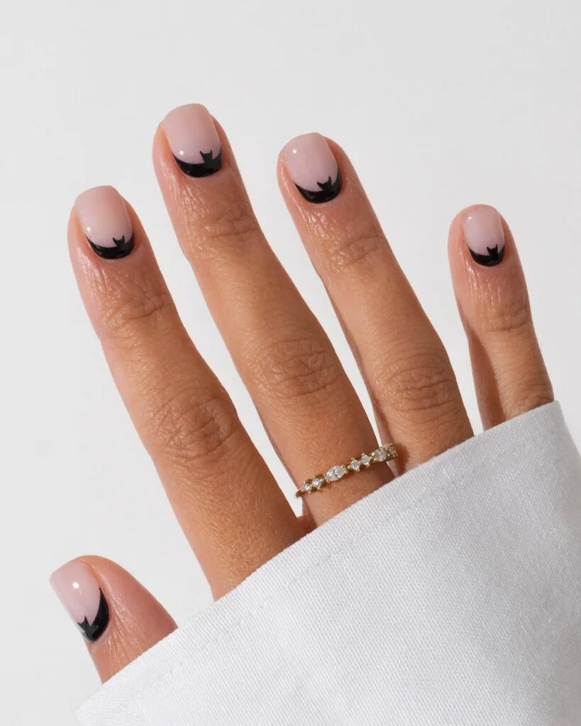 short Halloween nails with nude pink polish and black bat-wing reverse French details at the cuticles