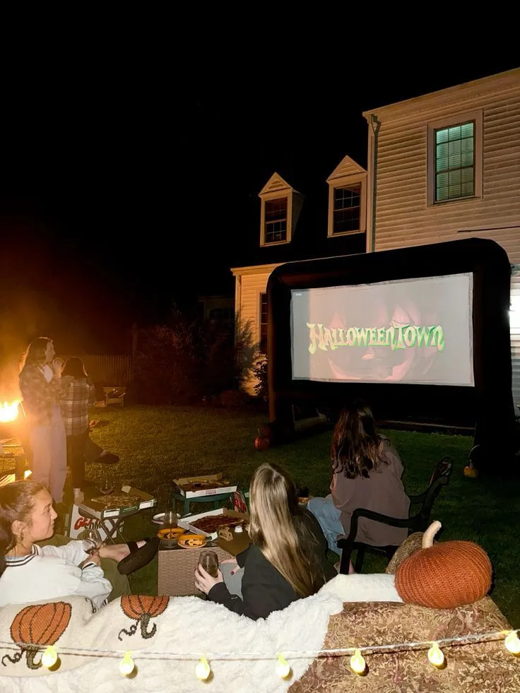 Outdoor Halloween movie night with a large screen, seated guests, blankets, string lights, and pumpkins
