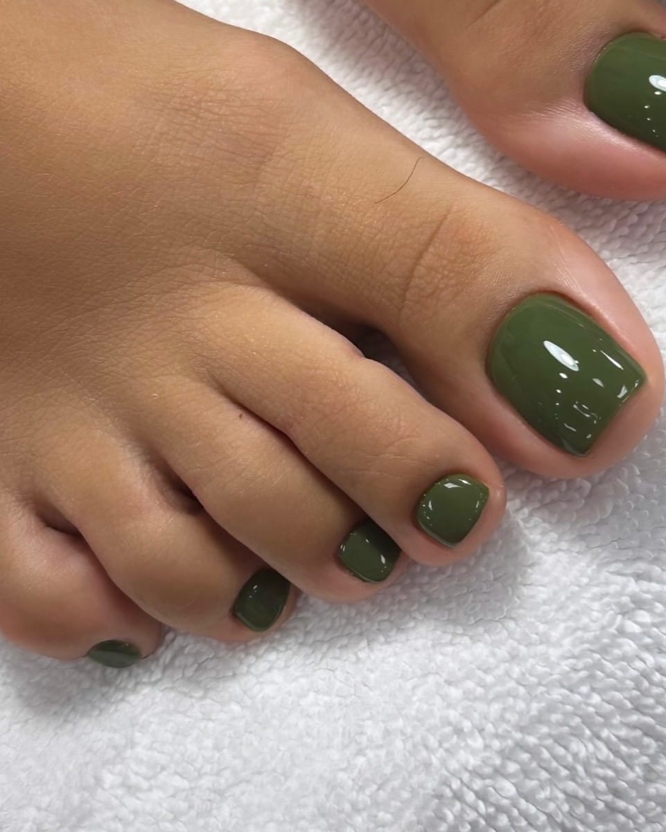 Dark olive green fall pedicure with glossy, softly squared toenails on a white towel.