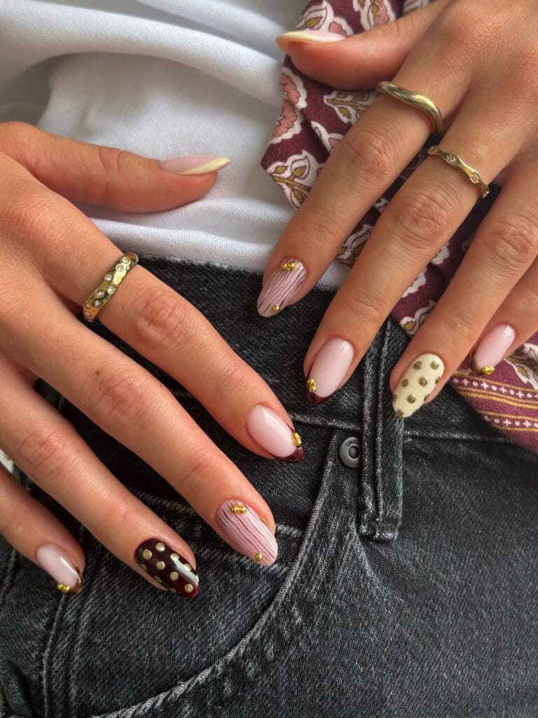 Pink fall nails with burgundy French tips, fine pinstripes, cream and brown accents, and tiny gold beads.
